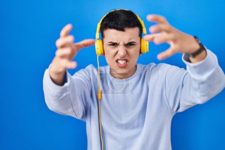 Foto de Persona no binaria escuchando música usando auriculares gritando frustrado con rabia, manos tratando de estrangular, gritando loco - Imagen libre de derechos
