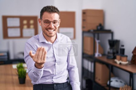 Foto de Joven hombre hispano en la oficina haciendo señas ven aquí gesto con la mano invitando a la bienvenida feliz y sonriente - Imagen libre de derechos