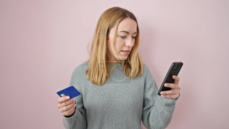 Foto de Mujer rubia joven de compras con teléfono inteligente y tarjeta de crédito sobre fondo rosa aislado - Imagen libre de derechos