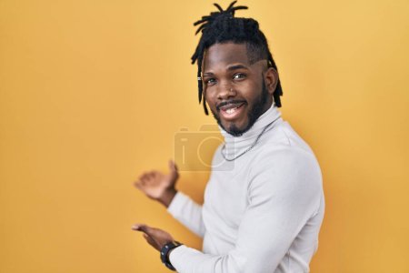 Foto de Hombre africano con rastas con jersey de cuello alto sobre fondo amarillo invitando a entrar sonriente natural con la mano abierta - Imagen libre de derechos