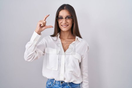 Photo for Young brunette woman wearing glasses smiling and confident gesturing with hand doing small size sign with fingers looking and the camera. measure concept. - Royalty Free Image