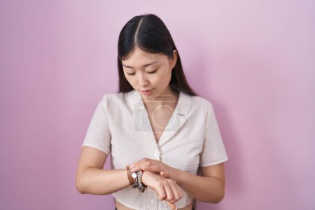Foto de Mujer joven china de pie sobre fondo rosa comprobar la hora en el reloj de pulsera, relajado y seguro - Imagen libre de derechos