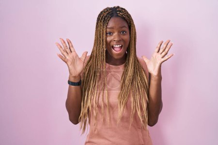 Foto de Mujer afroamericana con el pelo trenzado de pie sobre fondo rosa celebrando loco y sorprendido por el éxito con los brazos levantados y los ojos abiertos gritando emocionado. concepto ganador - Imagen libre de derechos