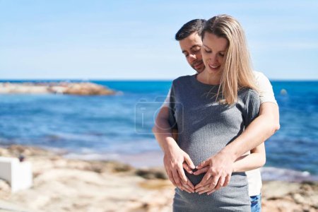 Foto de Hombre y mujer pareja abrazándose haciendo gesto de corazón en el vientre en la orilla del mar - Imagen libre de derechos
