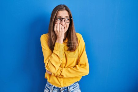 Photo for Young woman wearing glasses standing over blue background looking stressed and nervous with hands on mouth biting nails. anxiety problem. - Royalty Free Image