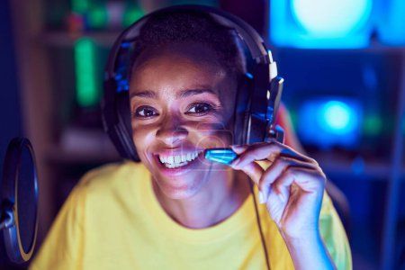 Téléchargez les photos : Femme afro-américaine streamer souriant confiant assis sur la table à la salle de jeux - en image libre de droit