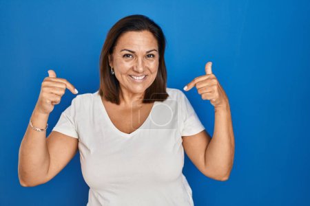 Téléchargez les photos : Femme mûre hispanique debout sur fond bleu regardant confiant avec le sourire sur le visage, se montrant du doigt fier et heureux. - en image libre de droit