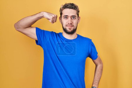 Foto de Hombre hispano con barba de pie sobre fondo amarillo persona fuerte mostrando el músculo del brazo, confiado y orgulloso de poder - Imagen libre de derechos