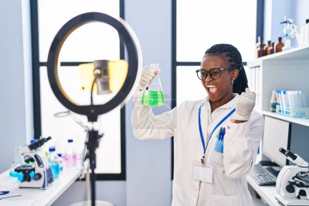 Foto de Mujer africana con trenzas trabajando en laboratorio científico haciendo tutorial con smartphone gritando orgullosa, celebrando la victoria y el éxito muy emocionada con el brazo levantado - Imagen libre de derechos