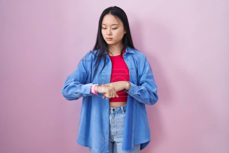 Photo for Young asian woman standing over pink background checking the time on wrist watch, relaxed and confident - Royalty Free Image