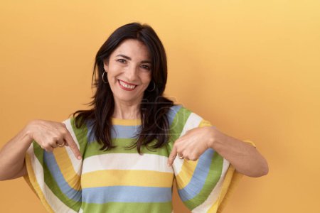 Foto de Middle age hispanic woman standing over yellow background looking confident with smile on face, pointing oneself with fingers proud and happy. - Imagen libre de derechos