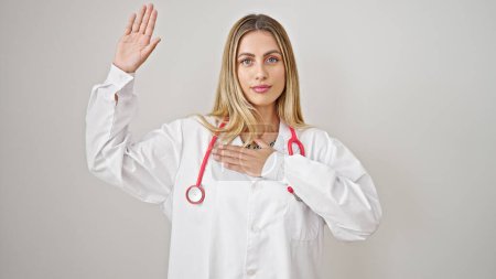 Foto de Joven doctora rubia haciendo un juramento con la mano en el pecho sobre fondo blanco aislado - Imagen libre de derechos