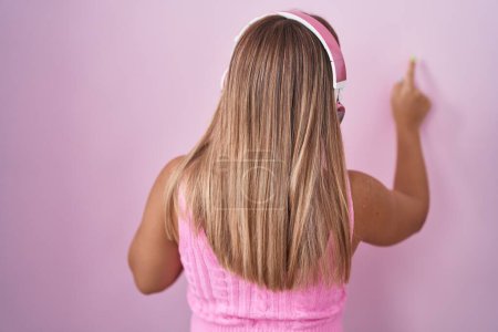 Foto de Mujer rubia joven escuchando música usando auriculares posando hacia atrás apuntando hacia adelante con la mano del dedo - Imagen libre de derechos