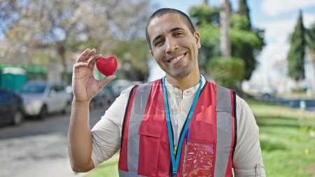 Foto de Joven hispano voluntario sonriendo sosteniendo el corazón rojo para la donación de sangre en el parque - Imagen libre de derechos