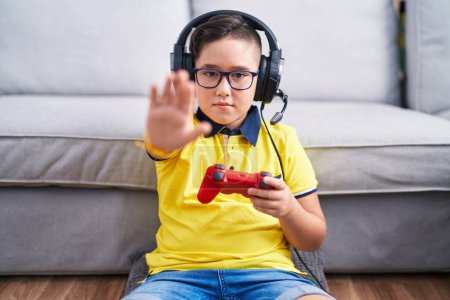 Photo for Young hispanic kid playing video game holding controller wearing headphones doing stop sing with palm of the hand. warning expression with negative and serious gesture on the face. - Royalty Free Image