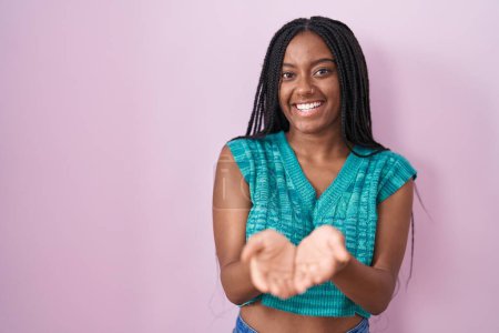 Foto de Joven afroamericano con trenzas de pie sobre fondo rosa sonriendo con las palmas de las manos juntas recibiendo o dando gesto. retención y protección - Imagen libre de derechos
