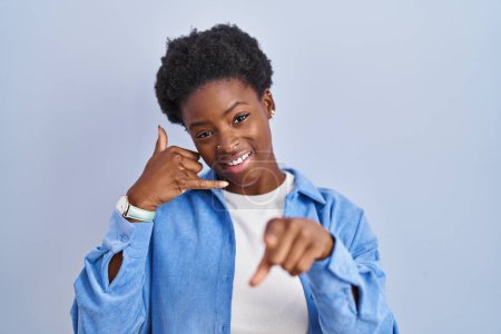 Téléchargez les photos : Femme afro-américaine debout sur fond bleu souriant faire parler au téléphone geste et vous pointant du doigt. Appelle-moi. - en image libre de droit