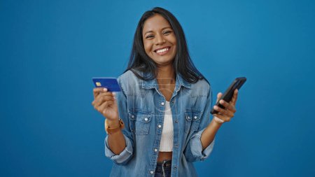Foto de Mujer afroamericana de compras con teléfono inteligente y tarjeta de crédito sobre fondo azul aislado - Imagen libre de derechos