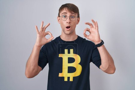 Téléchargez les photos : Homme blond caucasien portant Bitcoin t-shirt regardant surpris et choqué faire ok symbole d'approbation avec les doigts. expression folle - en image libre de droit