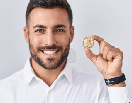 Foto de Hombre hispano guapo sosteniendo moneda criptomoneda litecoin mirando positiva y feliz de pie y sonriendo con una sonrisa confiada mostrando los dientes - Imagen libre de derechos