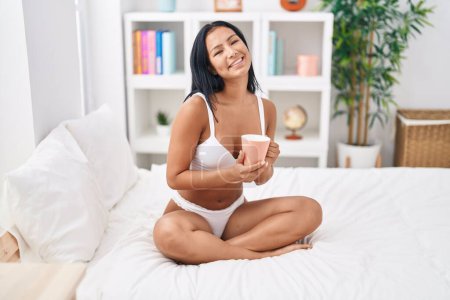 Téléchargez les photos : Young beautiful latin woman wearing lingerie drinking coffee at bedroom - en image libre de droit