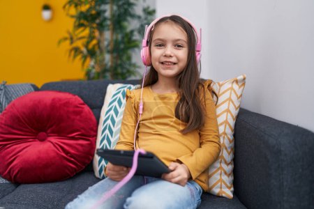 Téléchargez les photos : Adorable fille hispanique en utilisant touchpad et écouteurs assis sur le canapé à la maison - en image libre de droit
