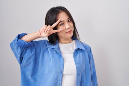 Téléchargez les photos : Jeune femme chinoise debout sur fond blanc faisant symbole de paix avec les doigts sur le visage, souriant gai montrant la victoire - en image libre de droit
