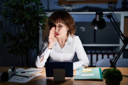 Téléchargez les photos : Femme d'âge moyen travaillant au bureau la nuit main dans la bouche racontant des rumeurs secrètes, chuchotant conversation malicieuse - en image libre de droit