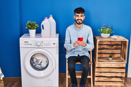 Photo for Young hispanic man using smartphone waiting for washing machine at laundry room - Royalty Free Image