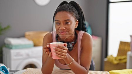 Téléchargez les photos : Femme afro-américaine sentant tasse de café appuyé sur la planche à repasser à la buanderie - en image libre de droit