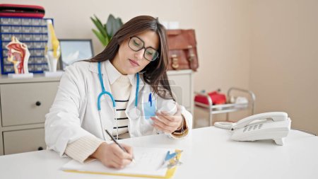 Foto de Joven hermosa mujer hispana médico sosteniendo pastillas botella de escritura en documento en la clínica - Imagen libre de derechos