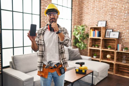 Photo for Young hispanic man with beard working at home renovation holding smartphone serious face thinking about question with hand on chin, thoughtful about confusing idea - Royalty Free Image