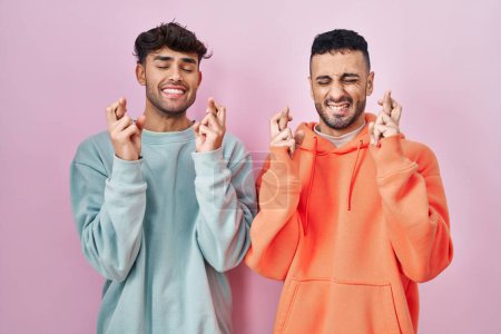 Téléchargez les photos : Jeune couple gay hispanique debout sur fond rose geste doigt croisé souriant avec espoir et les yeux fermés. chance et concept superstitieux. - en image libre de droit