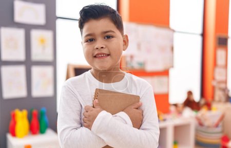 Téléchargez les photos : Adorable garçon hispanique debout avec cahier câlin à la maternelle - en image libre de droit