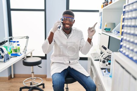 Photo for African american man working at scientist laboratory speaking on the phone smiling happy pointing with hand and finger to the side - Royalty Free Image