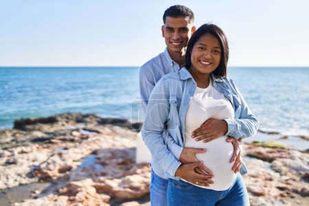 Foto de Joven pareja latina esperando un bebé abrazándose de pie en la playa - Imagen libre de derechos