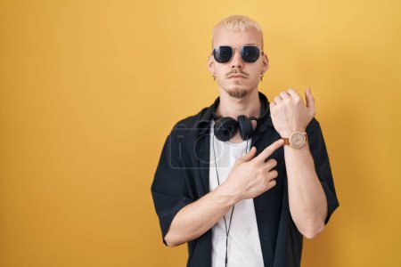Téléchargez les photos : Jeune homme caucasien portant des lunettes de soleil debout sur fond jaune à la hâte pointant vers regarder le temps, l'impatience, regardant la caméra avec une expression détendue - en image libre de droit