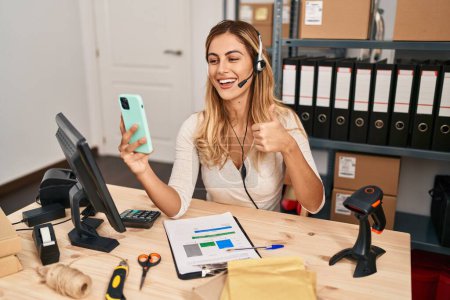 Photo for Young blonde woman working at small business ecommerce using headset smiling happy and positive, thumb up doing excellent and approval sign - Royalty Free Image