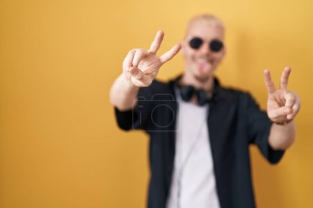 Foto de Joven hombre caucásico con gafas de sol de pie sobre fondo amarillo sonriendo con la lengua hacia fuera mostrando los dedos de ambas manos haciendo signo de victoria. número dos. - Imagen libre de derechos