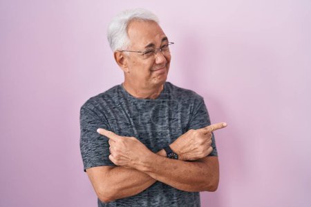 Téléchargez les photos : Homme d'âge moyen avec les cheveux gris debout sur fond rose pointant vers les deux côtés avec les doigts, direction différente en désaccord - en image libre de droit