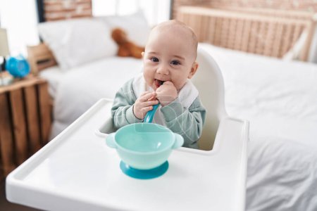 Photo for Adorable caucasian baby sitting on highchair sucking spoon at bedroom - Royalty Free Image