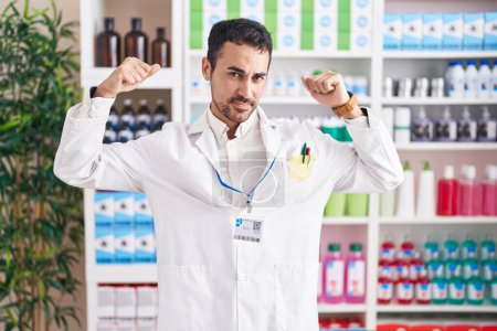 Téléchargez les photos : Un bel homme hispanique travaillant à la pharmacie montrant les muscles des bras souriant fier. concept de fitness. - en image libre de droit