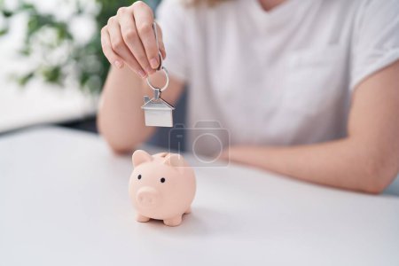 Photo for Young woman inserting house key on piggy bank at home - Royalty Free Image