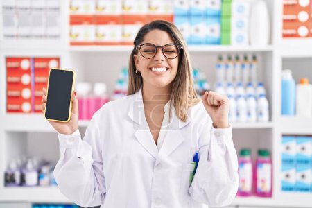 Téléchargez les photos : Femme blonde travaillant à la pharmacie drugstore montrant écran smartphone criant fier, célébrant la victoire et le succès très excité avec les bras levés - en image libre de droit