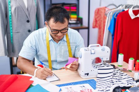 Téléchargez les photos : Jeune homme chinois tailleur écriture sur un bloc-notes tenant chiffon à la boutique de tailleur - en image libre de droit