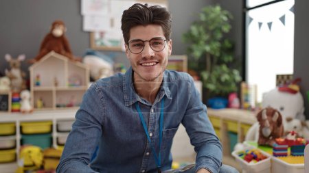 Téléchargez les photos : Jeune homme hispanique professeur préscolaire souriant confiant assis sur la table à la maternelle - en image libre de droit