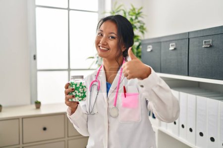 Téléchargez les photos : Jeune asiatique médecin femme tenant des pilules souriant heureux et positif, pouce vers le haut faire excellent et signe d'approbation - en image libre de droit