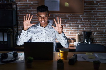 Photo for Young hispanic man working at the office at night showing and pointing up with fingers number nine while smiling confident and happy. - Royalty Free Image