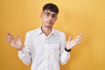 Téléchargez les photos : Young hispanic man standing over yellow background clueless and confused expression with arms and hands raised. doubt concept. - en image libre de droit