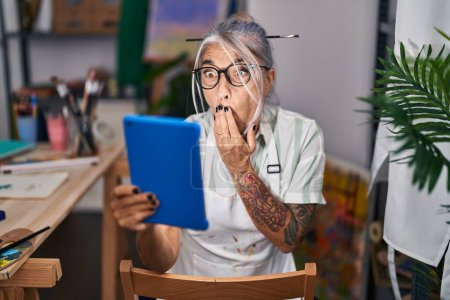 Photo for Middle age woman with grey hair sitting at art studio using tablet covering mouth with hand, shocked and afraid for mistake. surprised expression - Royalty Free Image